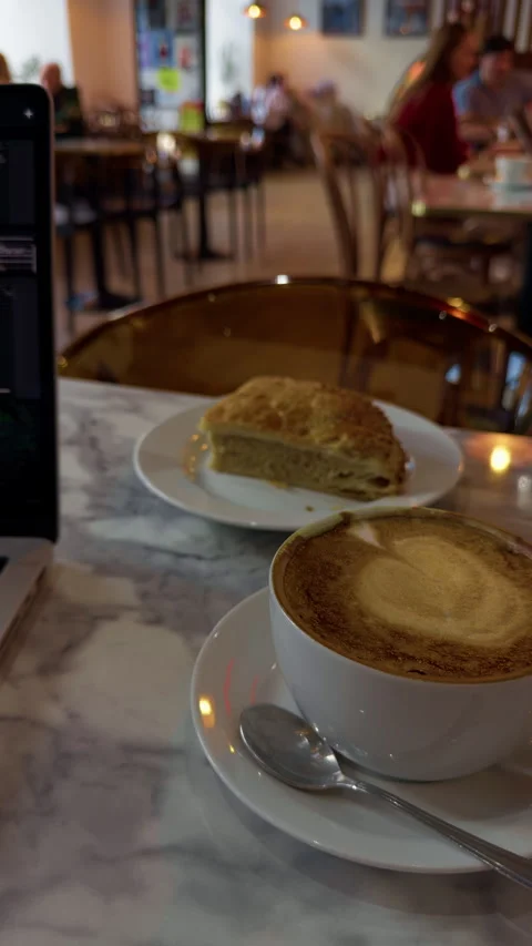 Cafe table with cappuccino, slice of cake, and softly blurred seating under warm Stock Footage 344967334