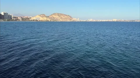 Calm blue sea with distant rocky hills and cityscape under clear sky in Alicante Stock Footage 294821315