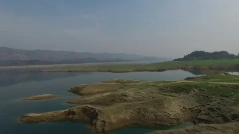 Calm lake waters reflecting rolling hills under a hazy blue sky Stockbeeldmateriaal 335270712