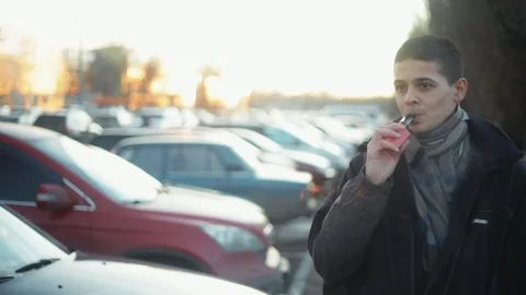 Calm man vaping e-cigarette between  cars on the parking zone near the mall, Видео 121483026