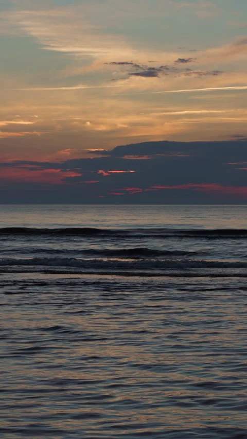 Calm ocean waves glow beneath colorful sunset sky, dark cloud band above horizon Stock Footage 345127829