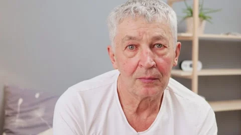 Calm relaxed gray haired man wearing white casual T-shirt sitting on bed looking 库存影片 265407707
