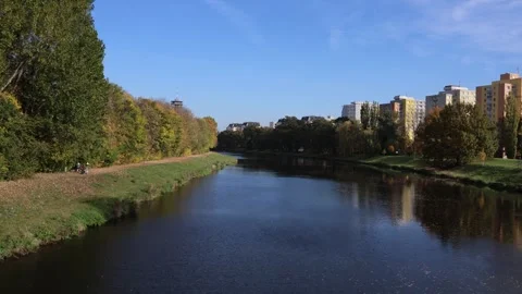 Calm river flows between autumn trees and city buildings under clear blue sky su Stock Footage 319841840