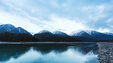 Calm river flows past snowy mountains under cloudy sky Stock Footage 333816713