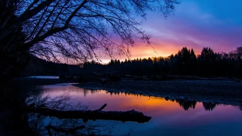 Calm river reflecting vibrant purple and orange sunset behind forest, wide shot Stock Footage 342396094