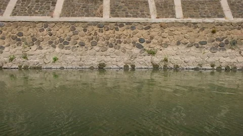 Calm water flowing alongside a structured stone retaining wall of a canal Video stock 347924073