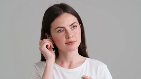 A calm woman wearing basic T-shirt is putting on wired earphones  動画素材 154946032
