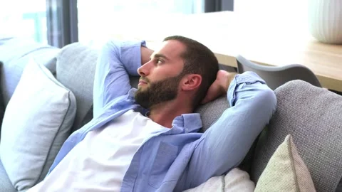 Calm young bearded man waking up from a nap on the sofa in his living room Stock Footage 344781926
