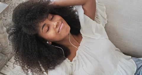 A calm young black lady using wireless headphones, reclining on a comfy sofa, Vídeo Stock 158806943