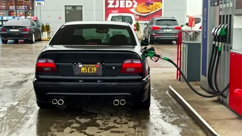 The camera approaches a classic black sedan being filled with fuel on a wet 库存影片 336179253