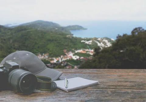 Camera with cap, notebook and pen on wooden table with mountain and sky backg Foto stock
