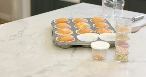 Camera capturing twelve golden cupcakes on countertop with sprinkles and whisk 스톡 동영상 335569808