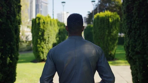 Camera follows behind a young man in a grey jacket as he walks through a city pa Stock Footage 117214480