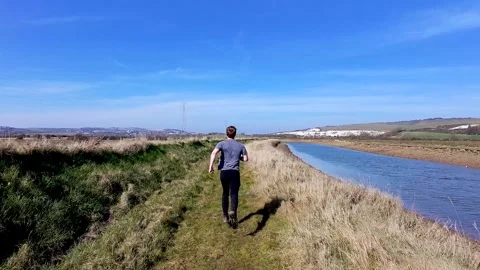 Camera follows man running along grassy trail next to river on spring sunny day Stock Footage 304298909