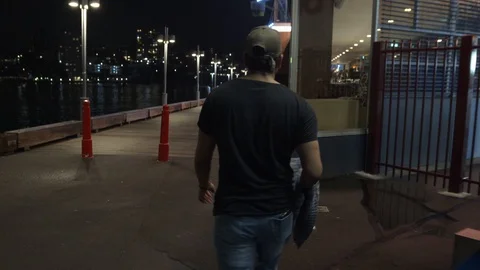 Camera Follows Man Walking on Dimly Lit Wooden Wharf at Night Stockbeeldmateriaal 107898808