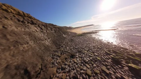 Camera moves along rugged coastline, showcasing sunlit water and pebbles Stock Footage 344563886