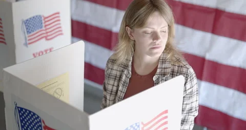 Camera moves gently on MS as young woman votes, US flag behind Stock Footage 274988816