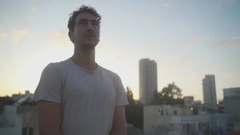 Camera moves towards a young serious man standing on a city roof at sunset Stock Footage 131900679