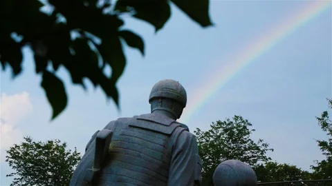 The camera pans past a rainbow filled sky and a stone soldier memorial. Stock Footage 112892787