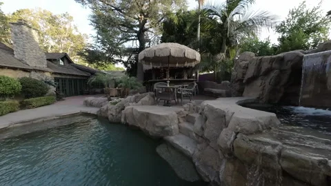 Camera passes palm fronds over a rock waterfall pool to a patio bar Stock Footage 345544158