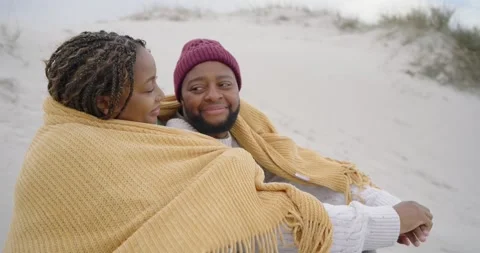 Camera pulling back, African-American couple wrapping mustard blanket on beach Stock Footage 340631261