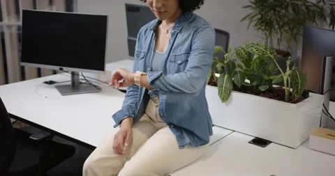 Camera pulling back African American woman sitting on desk checking watch Stock Footage 344672386