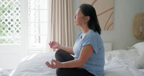Camera pulling back from knob, Senior Asian woman meditating on bed in tee Stockbeeldmateriaal 335269589
