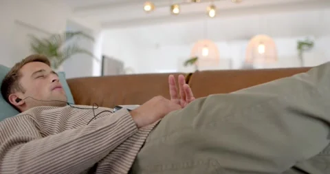Camera pulling back man reclining on sofa adjusting hands, listening with wired Stock Footage 344463205