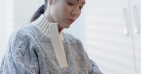 Camera pulling up, sweater-clad Asian woman, stapler on knee, lowering gaze, Stock Footage 339496244