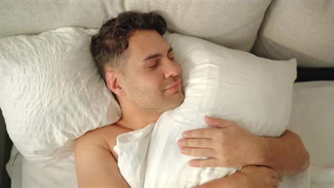 Camera pushes in on smiling man hugging white pillow as he opens his eyes. Stock Footage 346800286