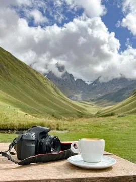 A camera rests on a table alongside a cup of coffee, with mountains Stock Photos