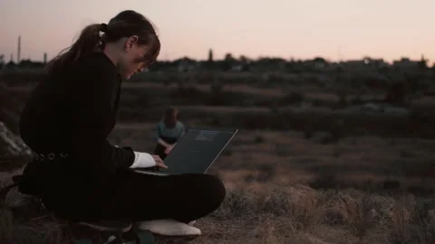 Camera rotates around modern styled girl working on laptop in a field in sunset Stock Footage 143271900