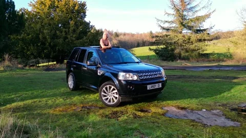 Camera Rotations Around Land Rover in Countryside with Elegant Woman in Sunroof Stock Footage 269564209