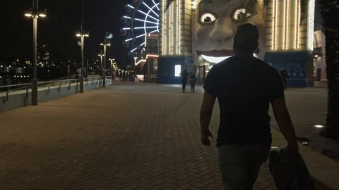 Camera Smoothly Follows Man Walking Towards Theme Park at Night Stock Footage 107898765