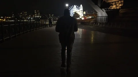 Camera Smoothly Follows Man Walking On Path Towards Theme Park at Night, Wide Stock Footage 107899195