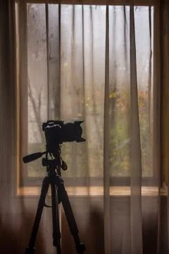 Camera on a tripod against the background of a window with a tulle curtain Stock Photos