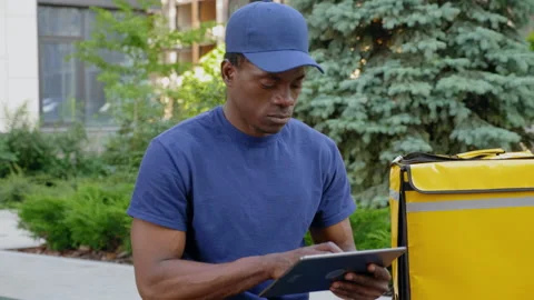 Camera zoom in afro-american man courier delivery sitting on bench uses tablet Stock Footage 157848320