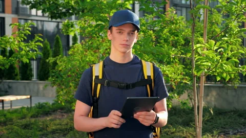 Camera zoom in young man courier food delivery with thermal backpack uses tablet Stock Footage 157848165