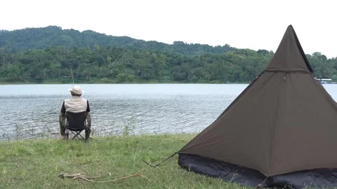 Camper fishing by a lakeside with a brown triangular tent in the background Stock Footage 327428614