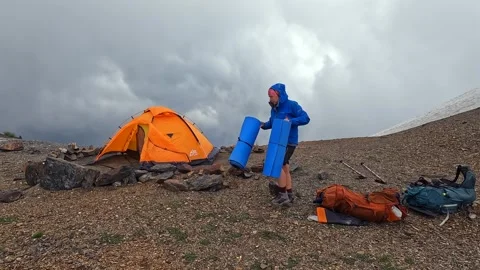 Camper packing gear wide view beside an orange tent on alpine campsite Stock Footage 343776098