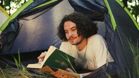 A camper reading a book inside a tent, illuminated by a flashlight, surrounded Stock Footage 285368207
