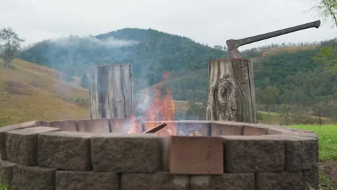 Campfire flames flicker in the foreground with an axe embedded in a chopping Stock Footage 344162656