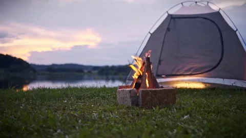 Campfire with glowing flames near camping tent by calm lake at sunset in Savci 動画素材 334042068