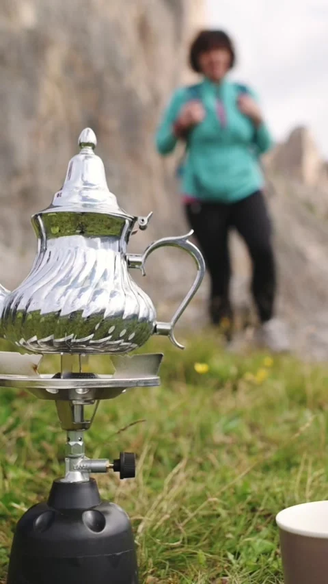Camping kettle and mug placed on grass with blurred woman and mountains in the Stockbeeldmateriaal 319063167