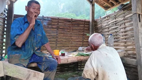 Candid Elderly Farmers Resting One Smoking and One Eating Inside Hut 스톡 동영상 335019478