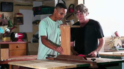 Candid senior carpenter guiding apprentice to use saw machine, slicing piec.. Stock Photos