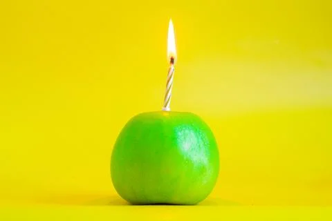 A candle burns in a green apple instead of a cake on a yellow background. Stock Photos