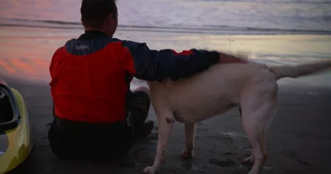 Canoeist sitting on the beach at sunset with his dog. Stock Footage 88373429