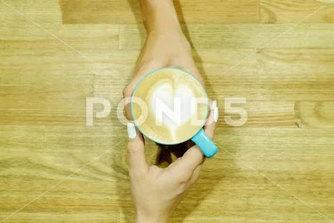 Cappuccino with heart froth between two hands on wooden table. Human pastime Stock Photos