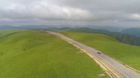 Car is going on asphalt road. Green hills and meadow, grey sky with clouds. A Stock Footage